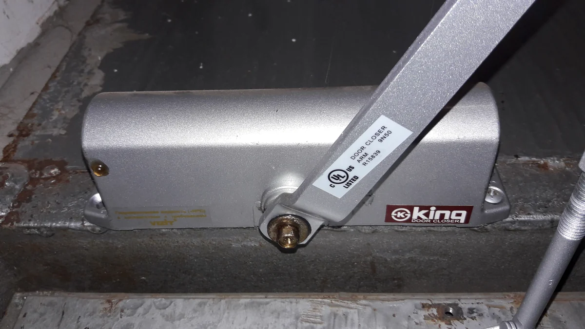 A silver surface mounted door closer screwed to a door with its jointed arm reaching up to the frame